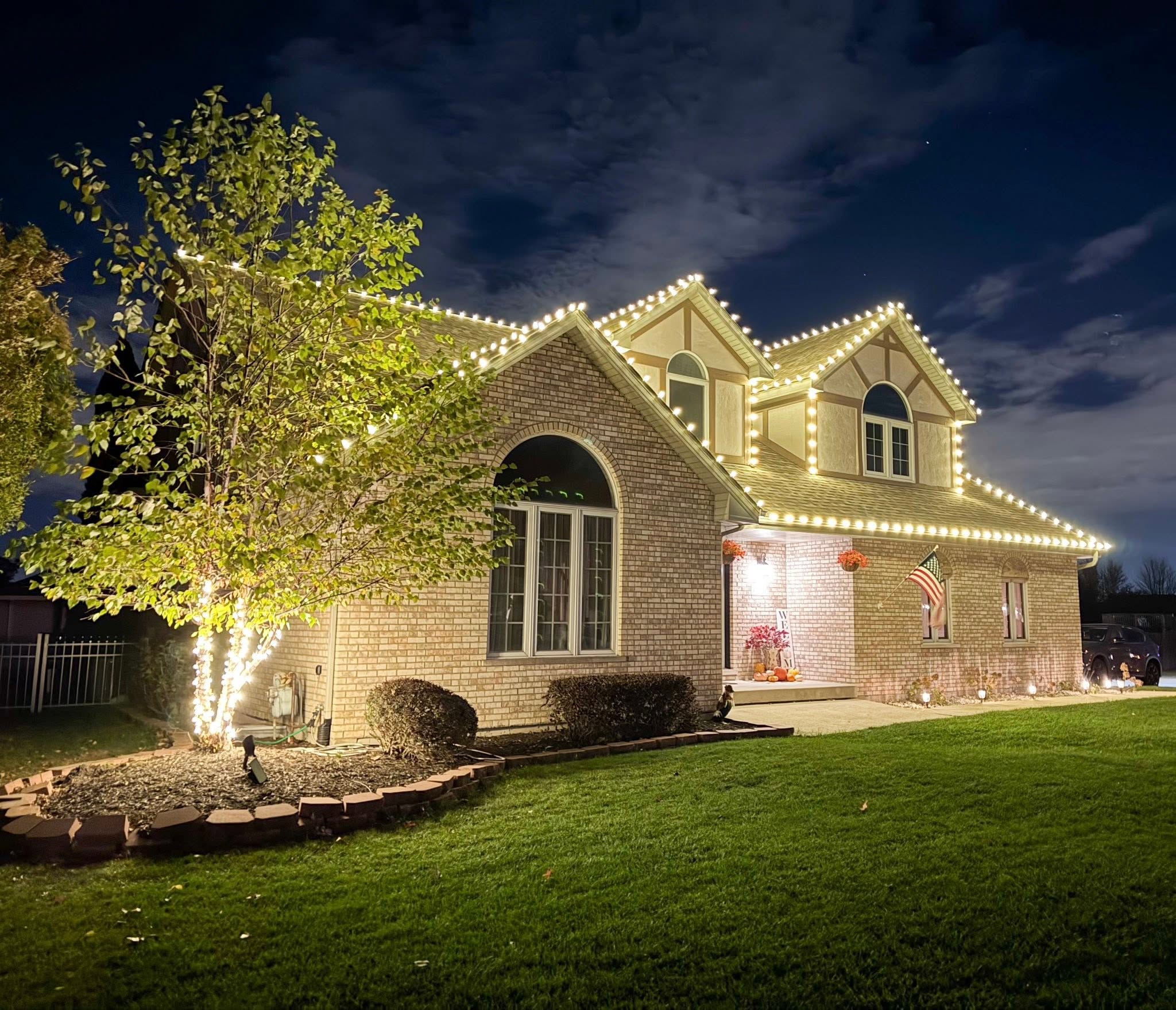 Brick home with warm white Christmas roofline lights and a wrapped front-yard tree